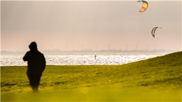 Die Silhouette einer Person ist links im Vordergrund zu sehen. Hinter einer Wiese fährt ein Kitesurfer auf der Nordsee.