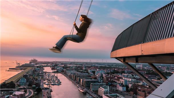 Eine Frau schaukelt im Sonnenuntergang über einer Stadt, sie schwingt frei in der Höhe. Rechts im Bild ist das Geländer einer Aussichtsplattform zu sehen. Im Hintergrund eine Stadt mit Hafen. Auch die Küste ist zu sehen.