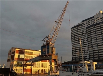 Historischer Hafenkran bei Sonnenuntergang in Bremerhaven, im Vordergrund ist ein Teil eines weißen Schiffs zu sehen, im Hintergrund ein modernes Hochhaus und ein Backsteingebäude mit großen Fenstern.