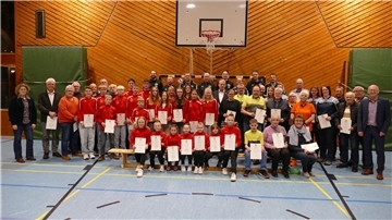 Eine große Gruppe von Menschen, darunter Kinder, Jugendliche und Erwachsene verschiedener Altersgruppen, posiert in einer Sporthalle für ein Gruppenfoto. Viele der Personen tragen rote Sportbekleidung, während andere in Alltagskleidung oder Vereinskleidung gekleidet sind. Sie halten Zertifikate oder Urkunden in den Händen. 