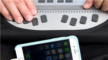 Eine Person bedient ein technisches Gerät und ist dabei mit einer Braillezeile und einem Smartphone mit Kopfhörern verbunden.