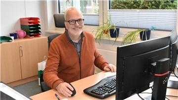 Ein älterer Mann mit einer brauen Jacke sitzt an einem Schreibtisch, auf dem ein Computer steht.