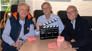 Drei Mitglieder des Kinoteams in der Amtsscheune sitzen lächelnd an einem Tisch. Einer hält eine Filmklappe mit der Aufschrift „Kinoteam in der Amtsscheune“, auf dem Tisch liegen Popcorntüten. Im Hintergrund sind Stuhlreihen zu sehen.