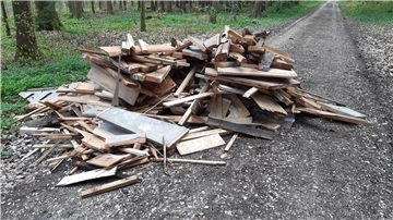 Ein großer Haufen aus Holzabfällen, darunter Bretter, Balken und andere Holzstücke, liegt am Rand eines unbefestigten Waldwegs. Im Hintergrund ist dichter Wald mit grünem Unterwuchs und hohen Bäumen zu sehen.