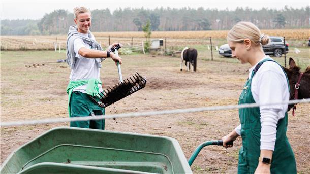 Alexandra Popp (links) steht bei ihrem Besuch auf einem Bauernhof in einem Tiergehege und schaufelt die Hinterlassenschaften in eine Schubkarre. Als gelernte Tierpflegerin ist sich die Fußball-Nationalspielerin dafür nicht zu fein.