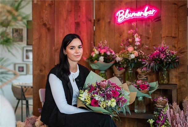 Eine Frau, die eine schwarze Hose und Weste über einem weißem Pullover trägt. Sie hält einen Blumenstrauß in der Hand. Hinter ihr sind noch weitere Blumensträuße zu sehen.