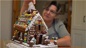 Landfrau Agnes Dahl sitzt an einem Tisch in ihrer Küche und stopft Watte in den Schornstein eines bunt geschmückten Lebkuchenhauses.