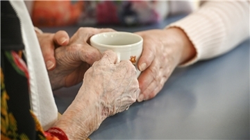 Eine Betreuungskraft hilft einer blinden Frau mit der Kaffeetasse in einem Seniorenheim.