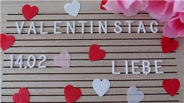 Eine Tafel auf der Valentinstag, 14.02. und Liebe geschrieben steht. Darum herum Konfettiherzen.