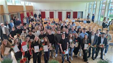 Ganz viele junge Männer und Frauen stehen in einem Raum mit Stühlen.