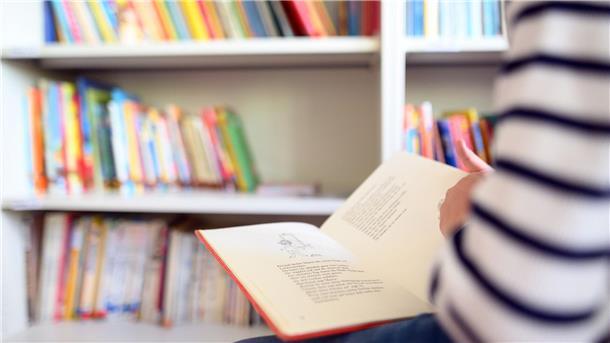 Ein Schüler liest ein Buch, im Hintergrund ist ein Regal mit vielen Büchern zu sehen.