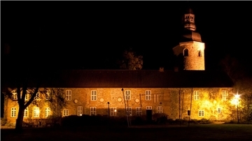 Ein langgestrecktes Gebäude und ein Kirchturm werden in der Dunkelheit angestrahlt.
