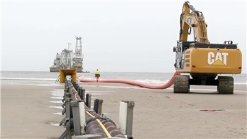 Am Strand von Norderney verschwindet ein Stromkabel im Meer. Im Hintergrund ein Verlegeschiff, von dem aus der Kabelstrang auf den Meeresboden hinabgelassen wird, bis hinaus zu einem Windpark.