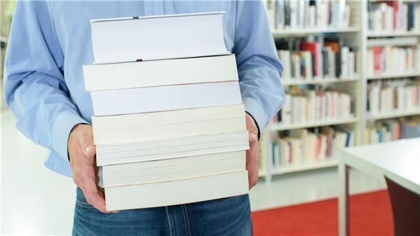 Zum Tag der Bibliotheken gibt es ein Blind Date, Fake News und Bilderbuchkino Ein Mann in einem blauen Hemd trägt einen Stapel Bücher.