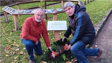 An der Gedenktafel wurden ein Gesteck und Blumen niedergelegt. Im Hintergrund sind die Tontafeln zu sehen, die an die 35 umgebrachten Babys erinnern.