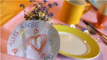 An einem Strauß Blumen auf einem gedeckten Frühstückstisch lehnt ein Papier mit der Aufschrift „Alles Liebe zum Muttertag“.
