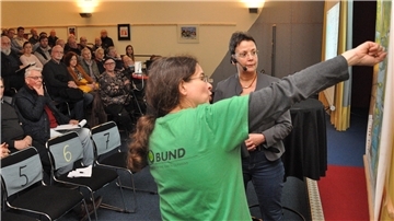 Zwei Frauen stehen im Güterschuppen in Nordenham vor Publikum an einer Landkarte.  Annette Chapligin vom BUND Wesermarsch nimmt jetzt Stellung zu den kritischen Anfragen der Union zu Nicht-Regierungs-Organisationen.