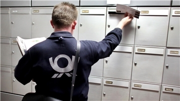 Ein Postzusteller steckt eine Sendung in einen Briefkasten. 