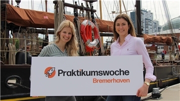 Zwei junge Frauen, Mitarbeiterinnen aus dem Standortmarketing der BIS Wirtschaftsförderung, halten vor der Kulisse des Neuen Hafens in Bremerhaven, ein Banner mit der Aufschrift "Praktikumswoche Bremerhaven"