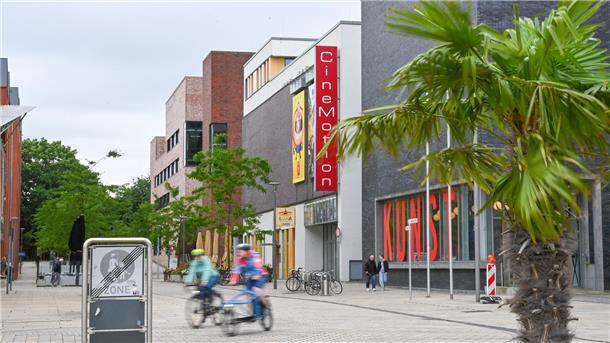 Blick auf das CineMotion-Kino in Bremerhaven mit rotem Fassadenschild. Im Vordergrund eine Palme und Radfahrende in Bewegung, rechts das Kunstmuseum mit großer „KUNST“-Beschriftung im Fenster.