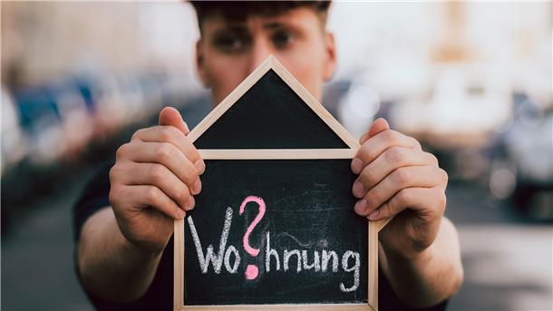 Ein Mann hält ein Schild mit einem Wohnungsgesuch in der Hand.