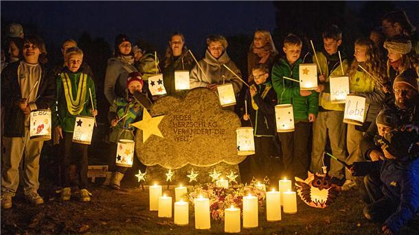 Eine Gruppe von Menschen hat sich in der Dunkelheit des Abends um einen Grabstein versammelt und hält leuchtende Laternen in der Hand.  Vor dem Grabstein leuchten Kerzen.  