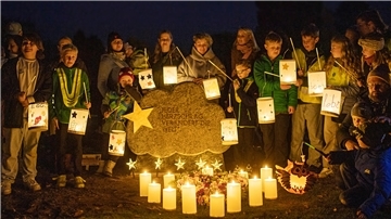 Eine Gruppe von Menschen hat sich in der Dunkelheit des Abends um einen Grabstein versammelt und hält leuchtende Laternen in der Hand.  Vor dem Grabstein leuchten Kerzen.  
