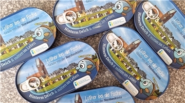 Mehrere Fischdosen liegen auf dem Boden. Die Dosen zeigen eine Kirche, Strandkörbe und den Slogan "Luther bei die Fische". Dazu werden schöne Grüße von der Nordsee gewünscht.