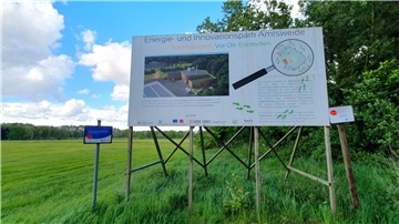 Ein großes Bauschild steht auf einer Wiese vor einem Waldrand. Es informiert über den „Energie- und Innovationspark Amtsweide“ in Geestland mit Text, Logos und einer Illustration des geplanten Geländes samt Solaranlagen und Gebäuden.
