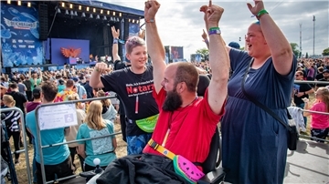 Eine Familie (Vater, Mutter, Sohn) reißt auf einem Festivalgelände vor der Bühne die Arme in die Luft. Sie befinden sich auf der Rolli-Tribüne. Der Vater nutzt einen Rollstuhl.