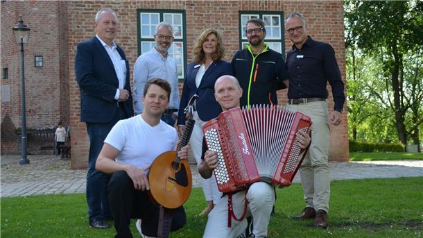 Sieben Personen posieren vor der Backsteinfassade der Burg Bederkesa. Zwei Musiker knien vorn mit Mandoline und Akkordeon, fünf weitere Personen stehen dahinter auf dem Rasen.