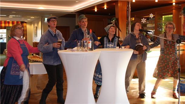 Eine Gruppe von sechs Frauen in rustikaler, ländlicher Kleidung steht gemeinsam an zwei weiß gedeckten Stehtischen und führt etwas vor.