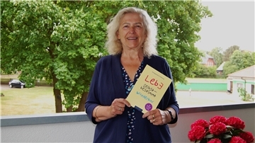 Eine ältere Frau mit blond-grauem Haar steht lächelnd auf einem Balkon vor grünem Baumbestand. Sie hält ein Buch mit dem Titel „Lebe selbstbestimmt wunderbar“ in den Händen. Auf dem Balkon stehen rote Blumen.