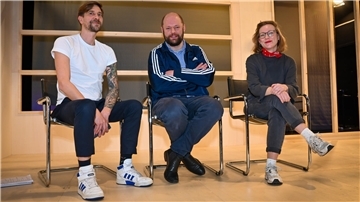 Eine Frau und zwei Männer sitzen auf Stühlen in einem Raum.