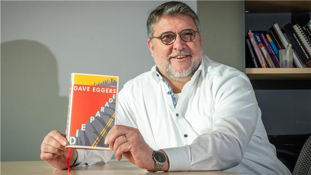 Ein Mann sitzt an einem Tisch und zeigt das Buch "Die Parade" in die Kamera.