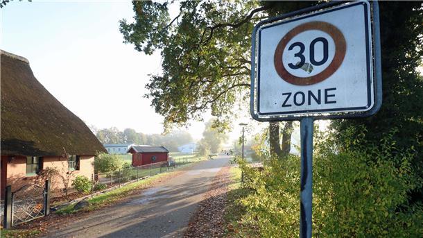 Eine Dorfstraße. Links ein Reetdachhaus, auf der rechten Straßenseite Gebüsch, ein Baum und ein Tempo-30-Schild