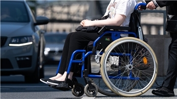 Person im Rollstuhl wird von einer Begleitperson auf einem Gehweg an einer Straße geschoben; im Hintergrund sind Autos im Stadtverkehr zu sehen.