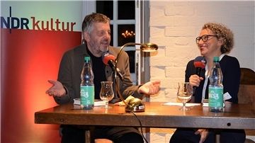 Zwei Personen sitzen bei einer Lesung an einem Pult und sprechen.