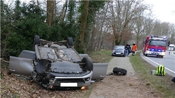 Ein Auto liegt nach einem schweren Unfall auf dem Dach. Im Hintergrund ist ein Fahrzeug der Feuerwehr zu sehen. 