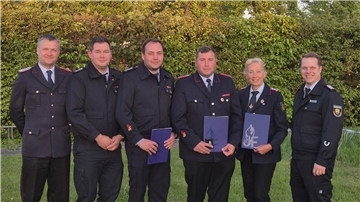 Sechs Mitglieder der Feuerwehr, teils in Uniform, stehen lächelnd nebeneinander. Drei von ihnen halten Ehrenurkunden und Auszeichnungen in den Händen.