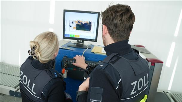 Ein Mann und eine Frau blicken auf ein Röntgengerät des Zolls.