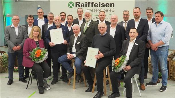 Eine Gruppe von Männern und Frauen posiert in einer Veranstaltungshalle vor einem Banner mit der Aufschrift „Raiffeisen Weser-Elbe eG“. Einige Personen in der ersten Reihe halten gerahmte Ehrenurkunden und Blumensträuße. 
