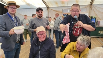 Drei Herren stehen beisammen. Einer hält einen Schweine-Schwanz in der Hand und hält diesen einer Dame vor die Nase. 