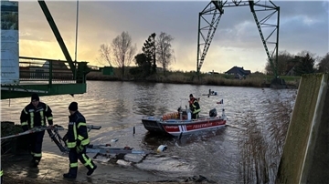 Ein Feuerwehrboot auf einem Fluss, besetzt mit Feuerwehrleuten in Einsatzkleidung, wird am Ufer von zwei weiteren Feuerwehrleuten vorbereitet oder gesichert.