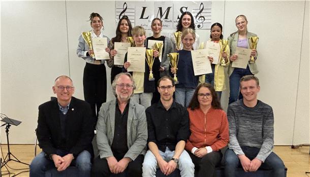 Acht junge Musikerinnen und Musiker stehen mit Urkunden und Pokalen in den Händen. Vor ihnen sitzen fünf Erwachsene. Im Hintergrund hängt das Logo der Loxstedter Musikschule.