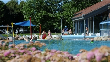 Ein Freibad ist zu sehen. Im Hintergrund befinden sich Menschen in Badekleidung, die planschen und spielen. 