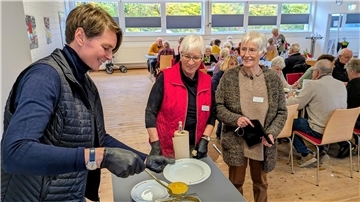 Menschen sitzen in einem Raum an Tischen. Im Vordergrund wird von einer Frau mit einer Kelle Suppe auf einen Teller gelöffelt.