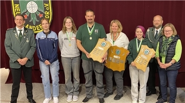 Auf dem Foto stehen acht Personen nebeneinander vor einer weinroten Bühne mit dem Vereinswappen des Schützenvereins Neuenlandermoor. Mehrere tragen grüne Schützenuniformen oder Vereinskleidung, einige haben Medaillen um den Hals. Drei Personen in der Mitte halten große Holztafeln mit eingravierten Namensschildern, die als Siegerplaketten dienen. Alle blicken in die Kamera und lächeln.