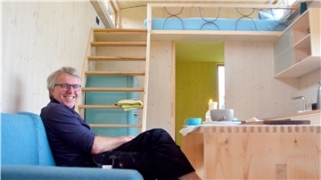 Bernd Klöpper auf dem Sofa in seinem Tiny House. Im Hintergrund ist die Treppe zum Hochbett zu sehen. Darunter steht der Kühlschrank.