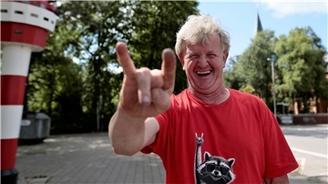 Ein Mann im roten T-Shirt lacht und hält die Hand zur Rockerpose „Pommesgabel“ hoch. 